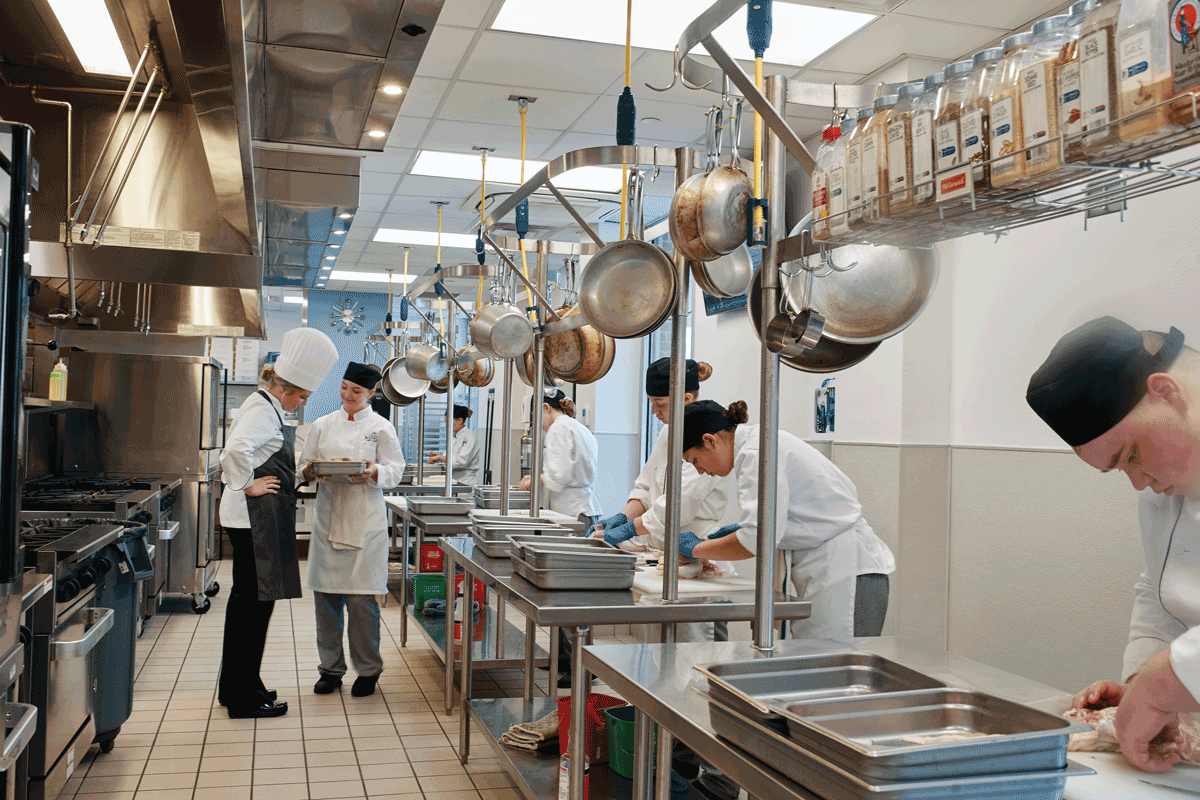 A commercial instructional kitchen with students bent over tables deconstructing chicken. The head chef looks at a student's work.