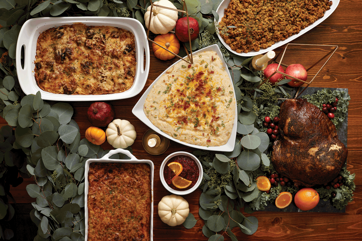Dark wooden table adorned with eucalyptus garland, small white pumpkins, a pomegranate, candles and a metallic geometric decor item, a turkey, mashed potatoes, casseroles, cranberry sauce.