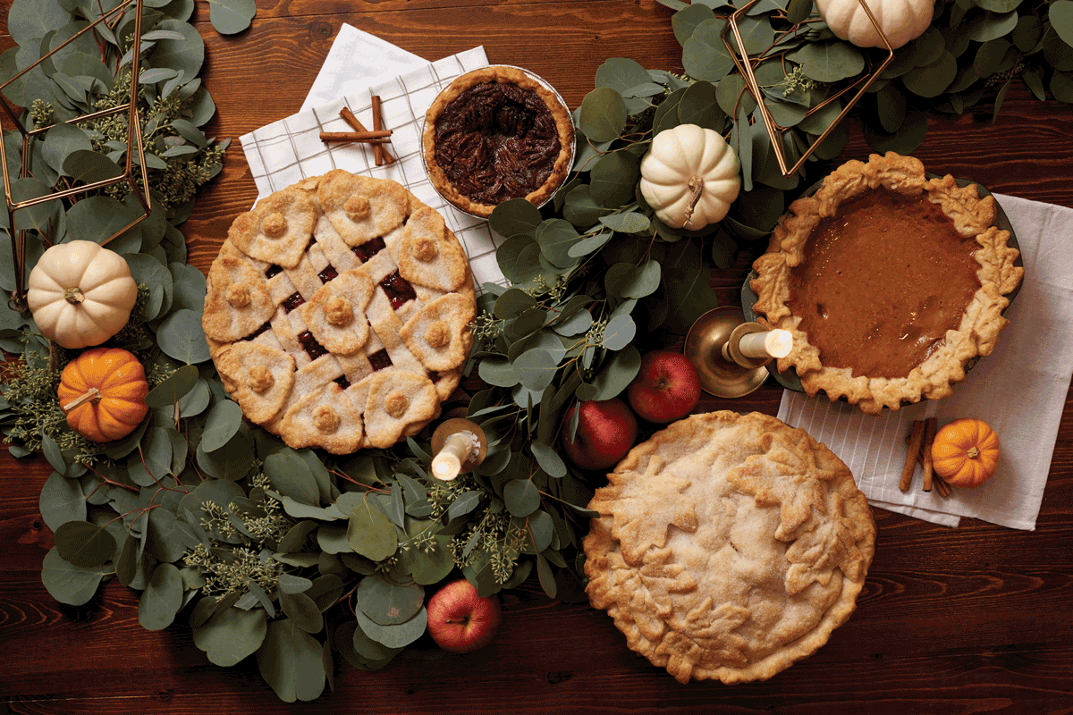 Dark wooden table adorned with eucalyptus garland, small white pumpkins, a pomegranate, candles and a metallic geometric decor items, cherry pie, pecan pie, apple pie and pumpkin pie.